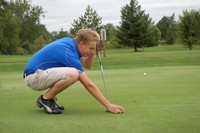 A person in a blue shirt and khaki shorts is crouching on a green golf course, lining up a putt. A yellow golf ball is nearby, and trees are visible in the background under an overcast sky.