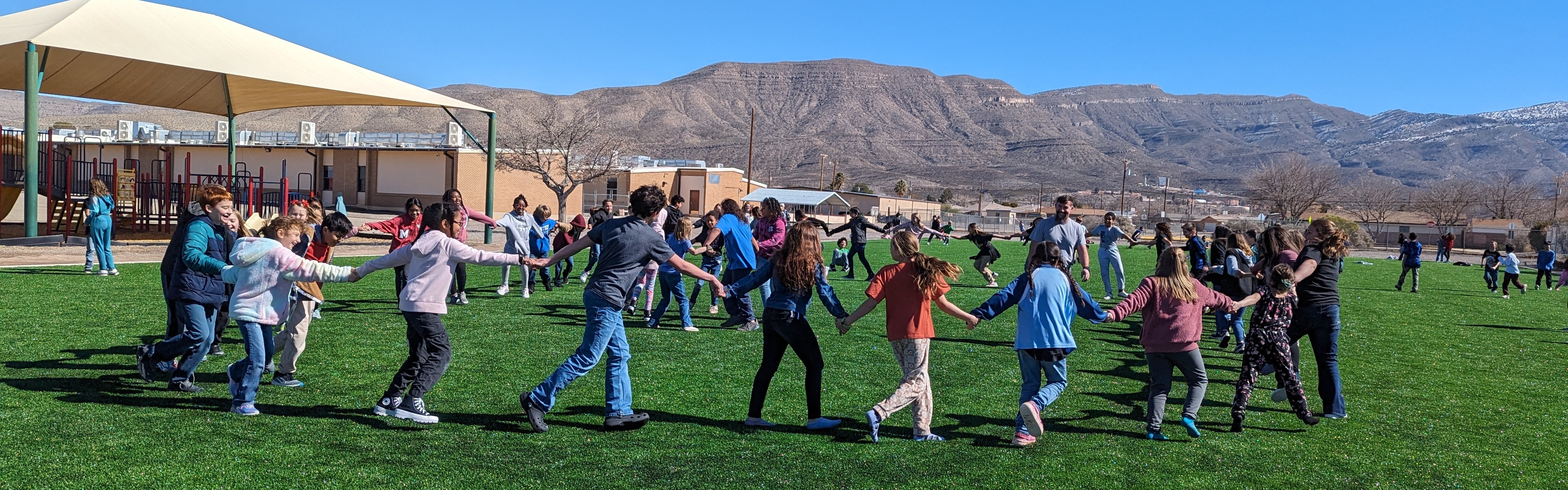 Students holding hands in a circle on green turf field with mountains in the background