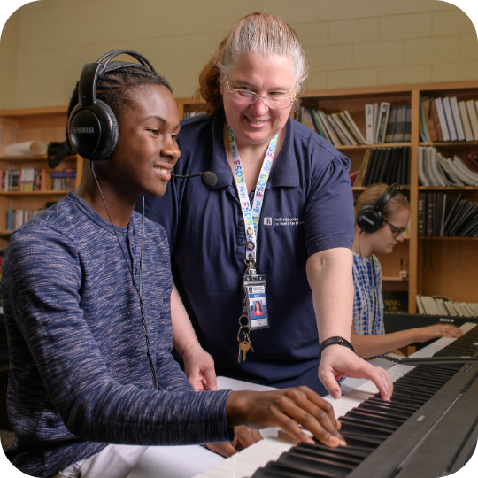 Blind High School boy playing the piano while a teacher helps him.