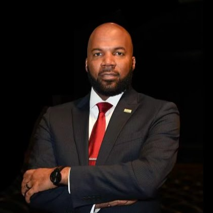 Man in a dark suit with a red tie, standing with arms folded, and wearing a watch.