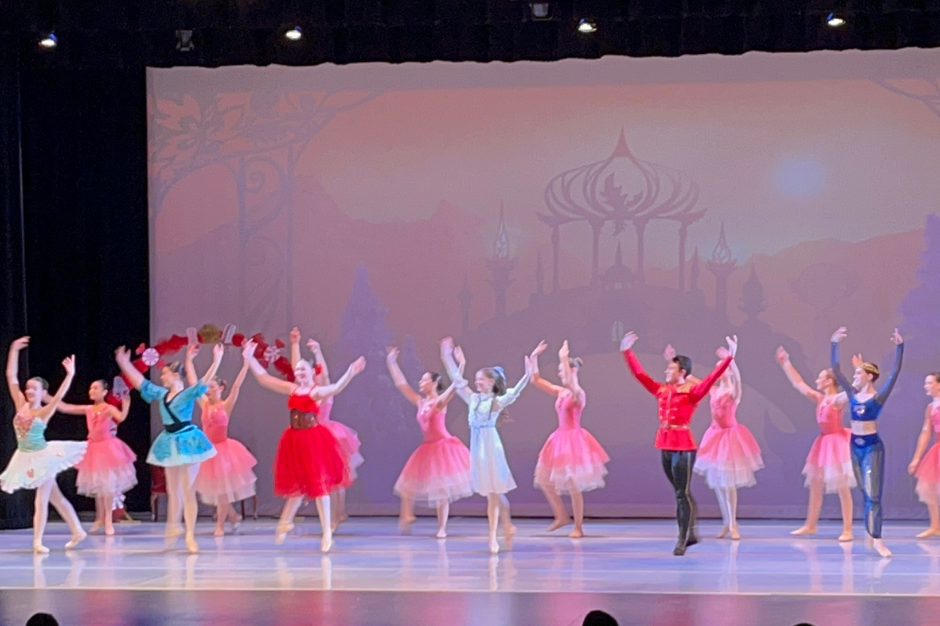 A ballet troupe in vibrant costumes performs on stage with an animated backdrop, under stage lights.