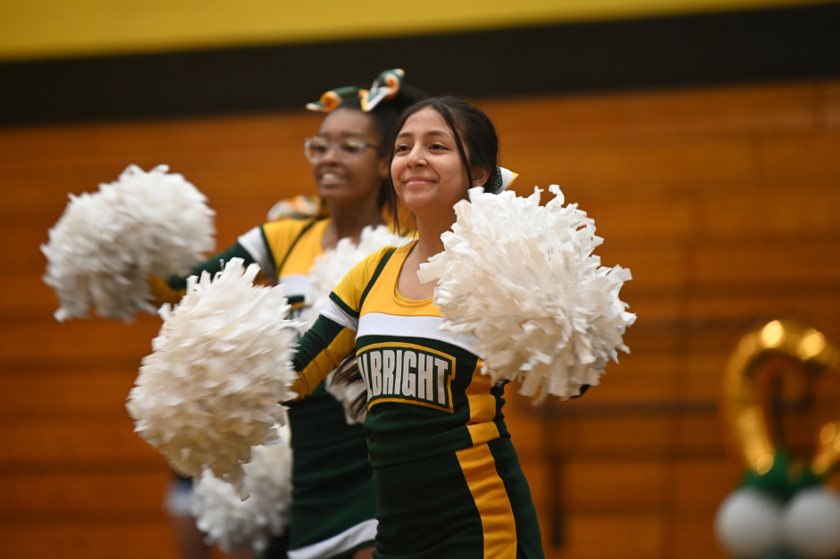 Two cheerleaders in green and yellow uniforms with white pom-poms smile. Behind them is a wooden wall.