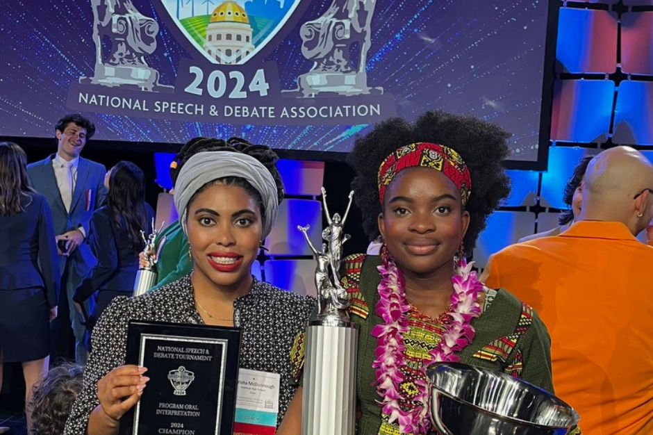 Two women stand holding trophies, one with a plaque, smiling. A large screen reads "2024 National Speech & Debate Association".