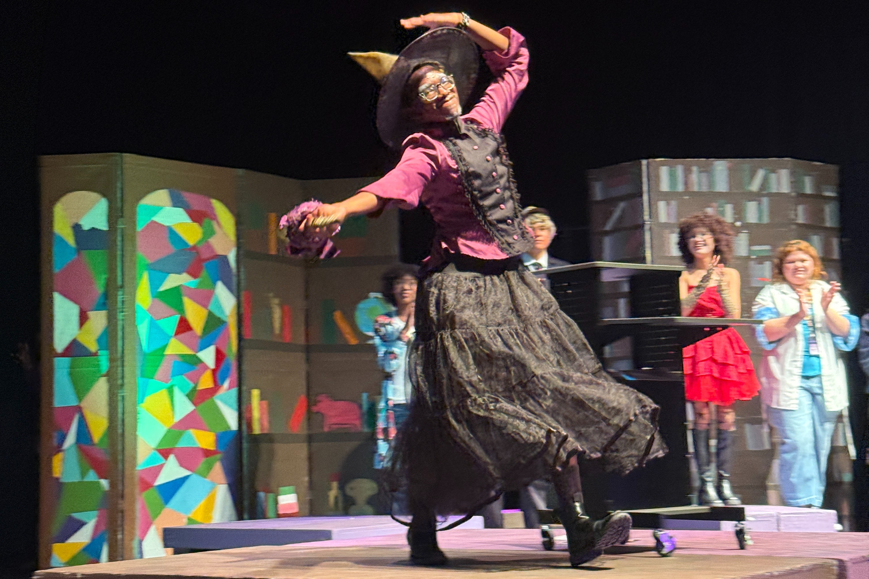 A performer on a raised stage with colorful set pieces behind them. Other people are in the background.