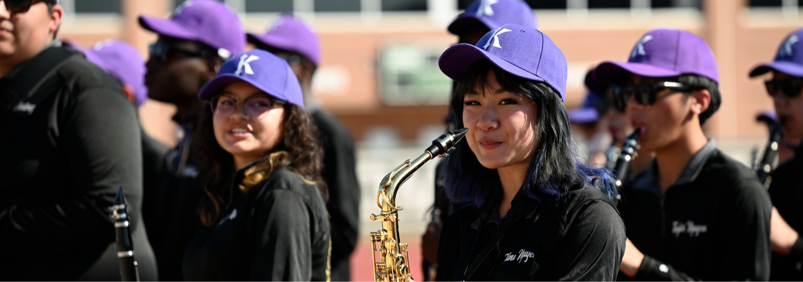 Musicians stand in a row wearing purple caps. A woman plays a saxophone. A building is in the background.