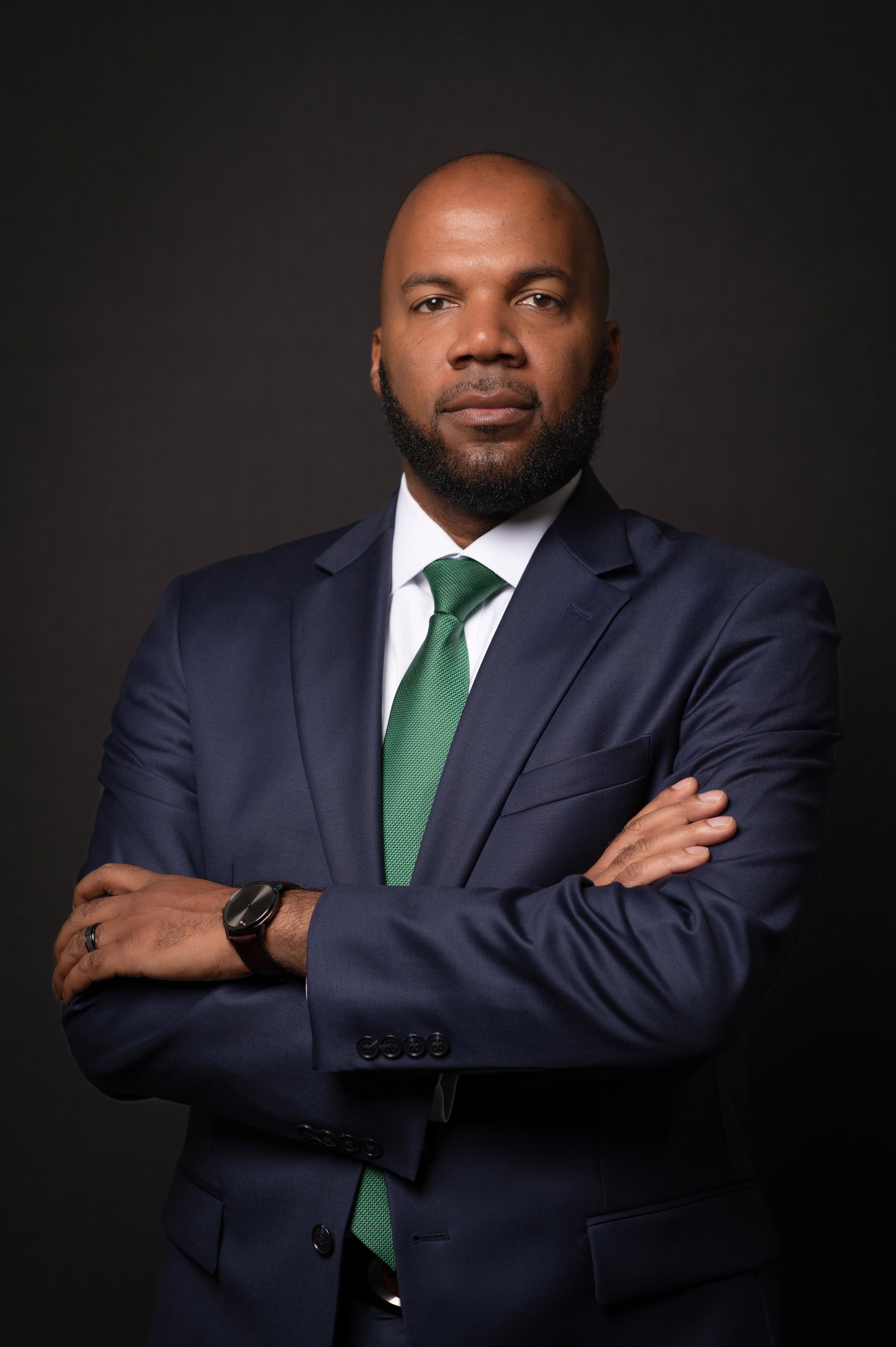 Man in business attire poses with folded arms against a dark background. He wears a dark blue suit and green tie.