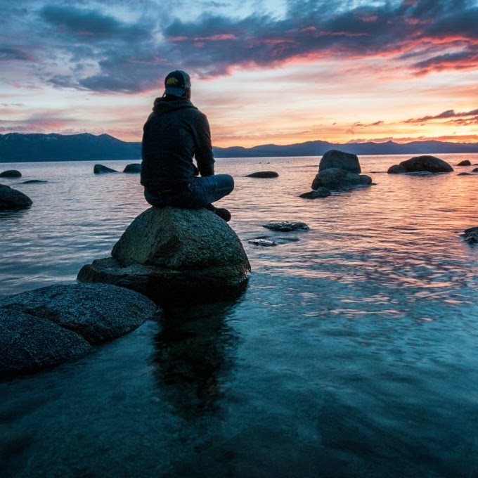 Person sitting on a rock by the water, enjoying the tranquil beauty of the sunset over the lake.