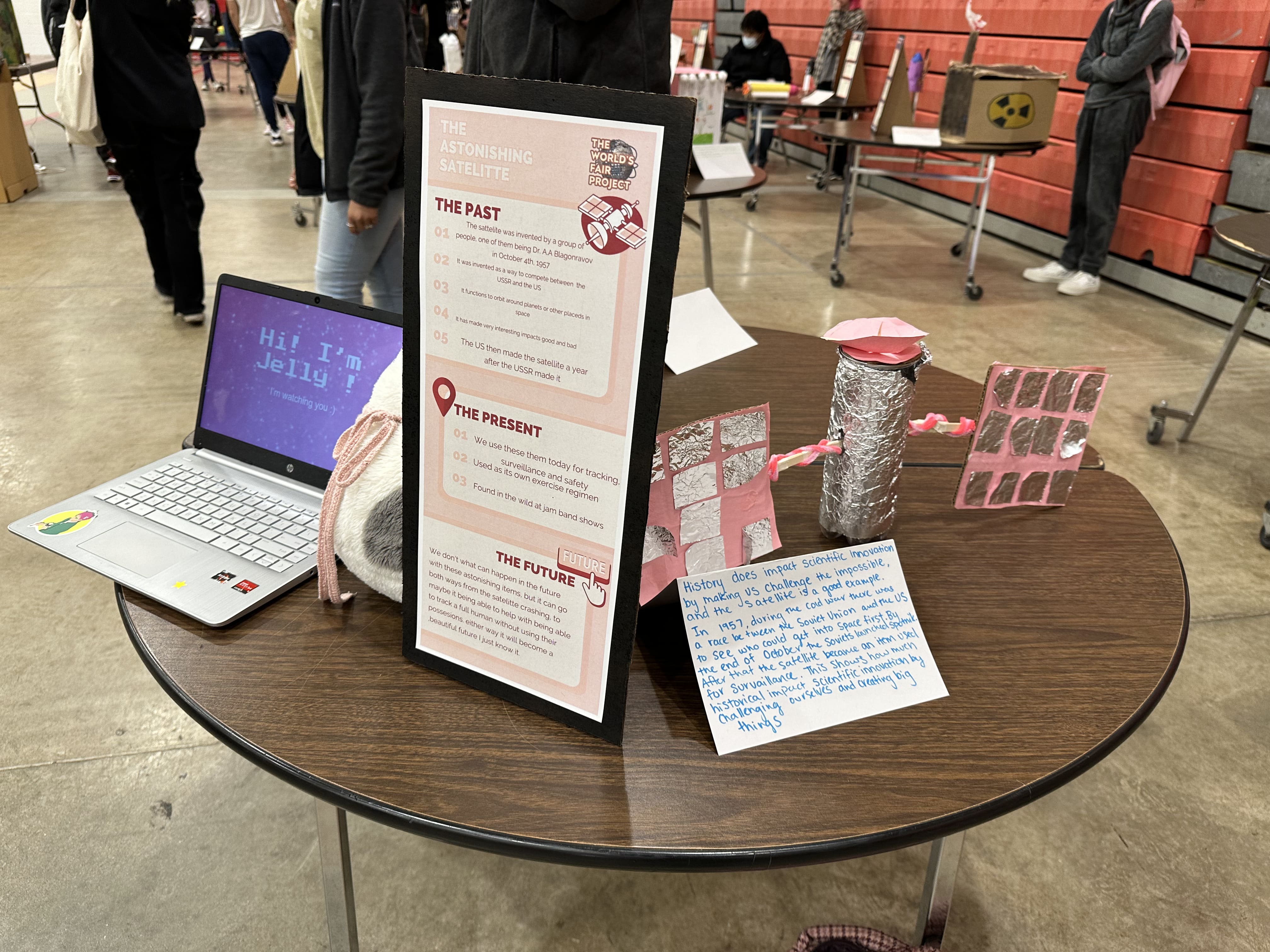 Display of student work for World's Fair Project. There is a laptop with a presentation on the screen that says, "Hi, I'm Jelly!" , There's a  thin  tall poster in pink that shows the past, present, and future of satellites, and on the  right on the table there's a small student made model of a satellite.