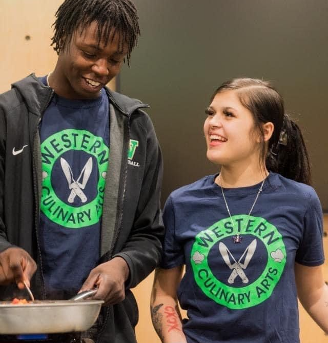 two students cooking food over a hot plate smiling and talking to each other