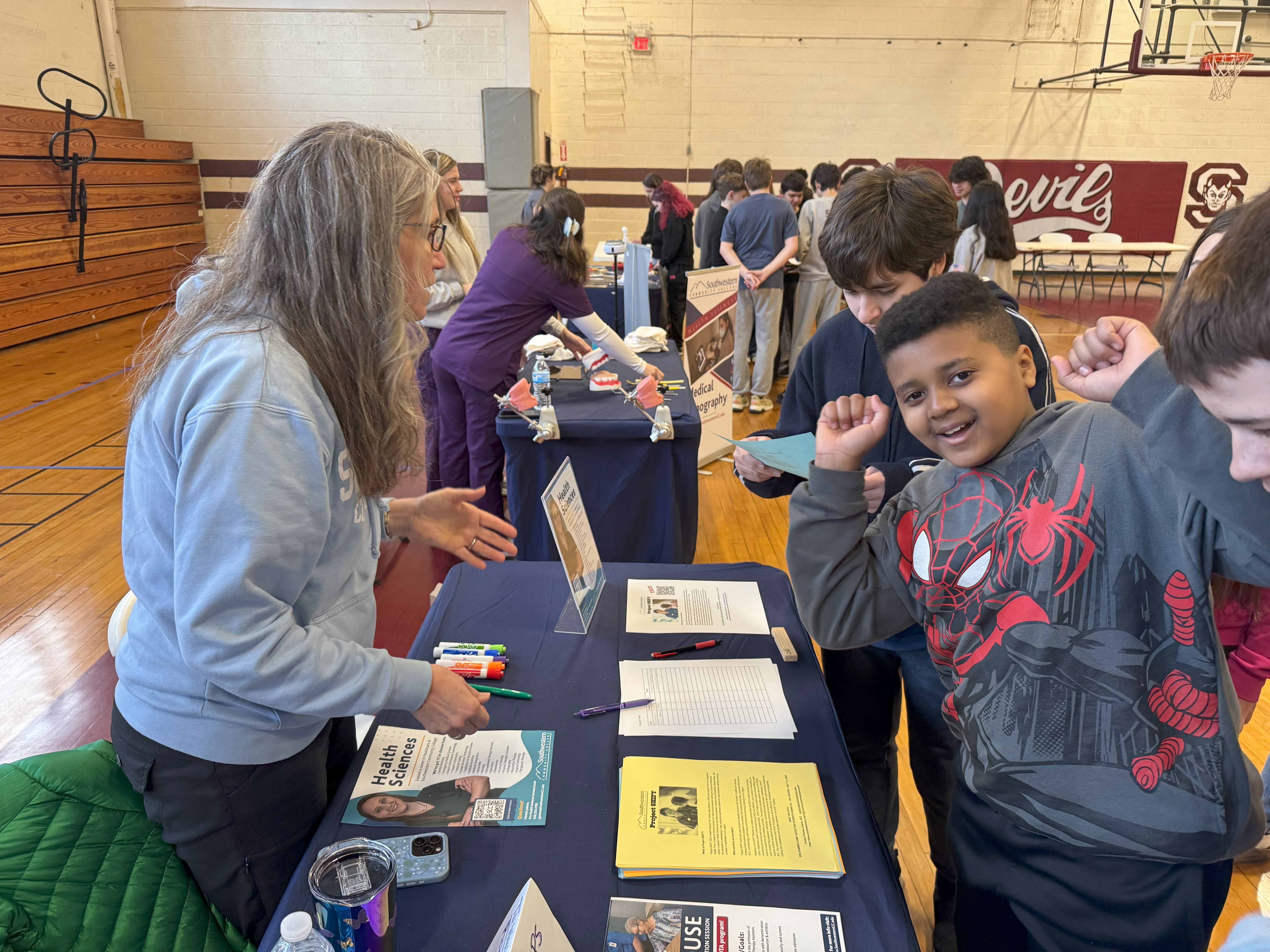 People interacting at an informational event in a gymnasium, featuring tables with educational materials.