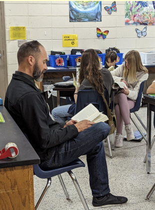 teacher seated reading a book at a classroom