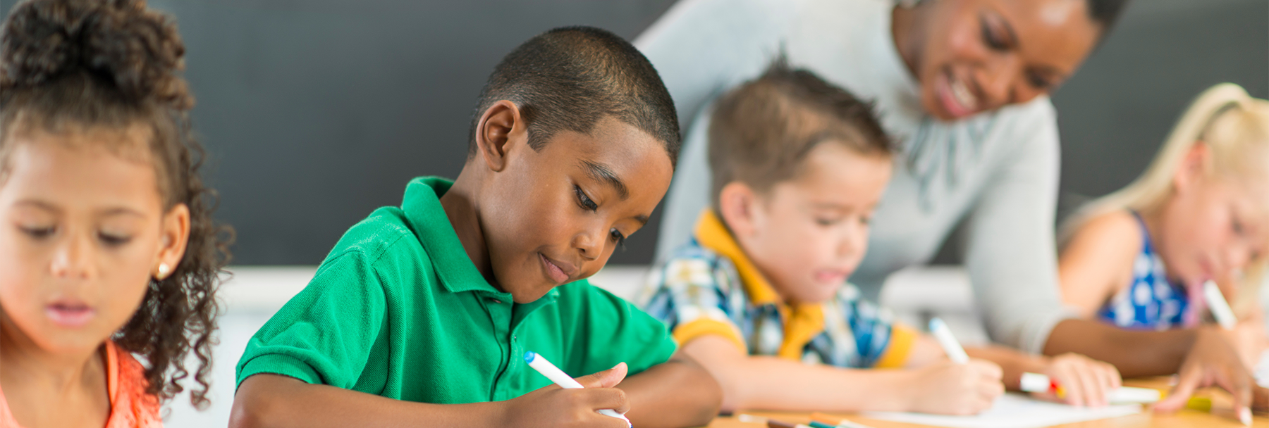 Boy coloring sitting at a desk, blurry group of students and teacher coloring at the background