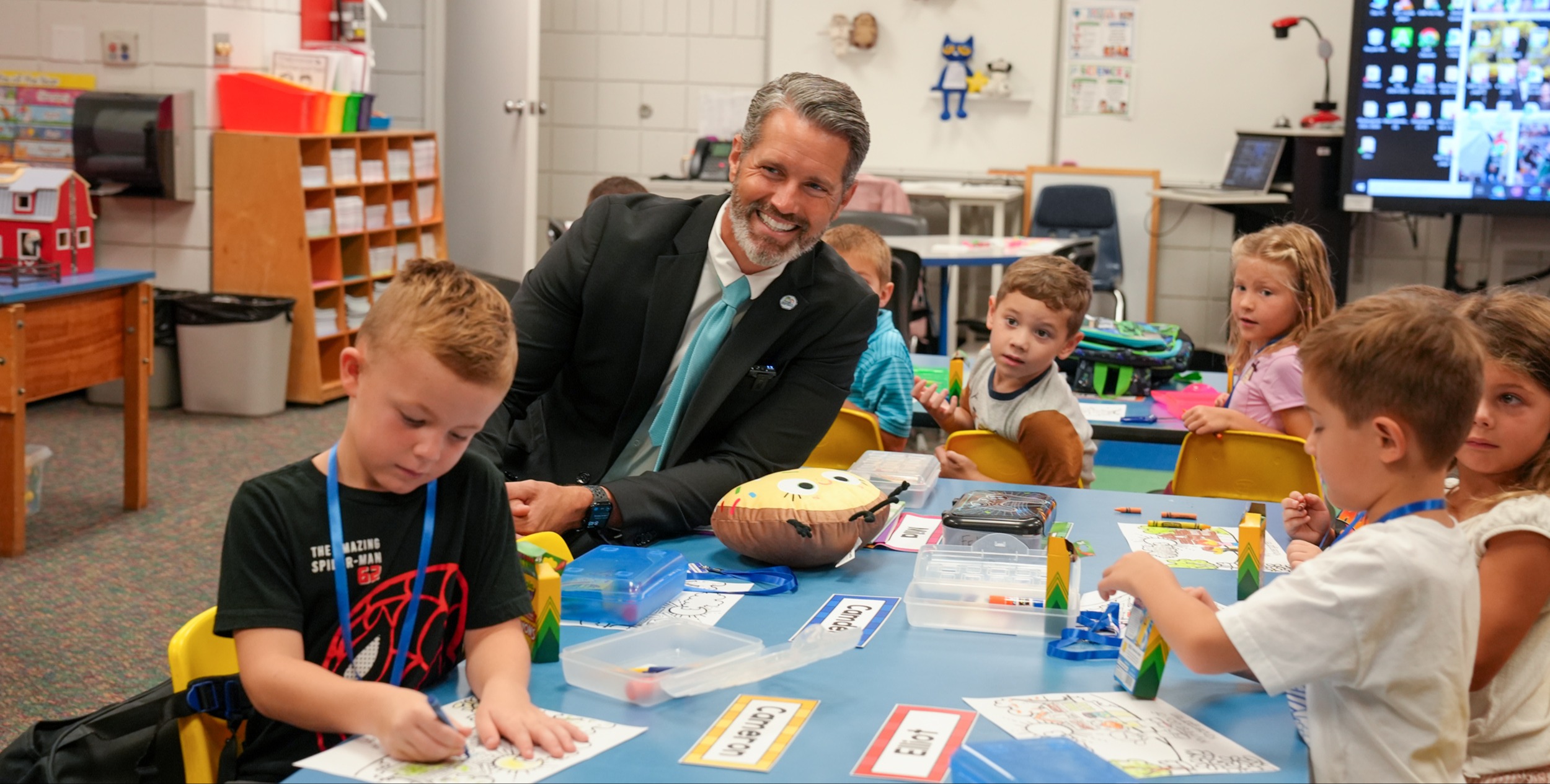 A man in a suit sits at a table with children. They are doing crafts at a desk.