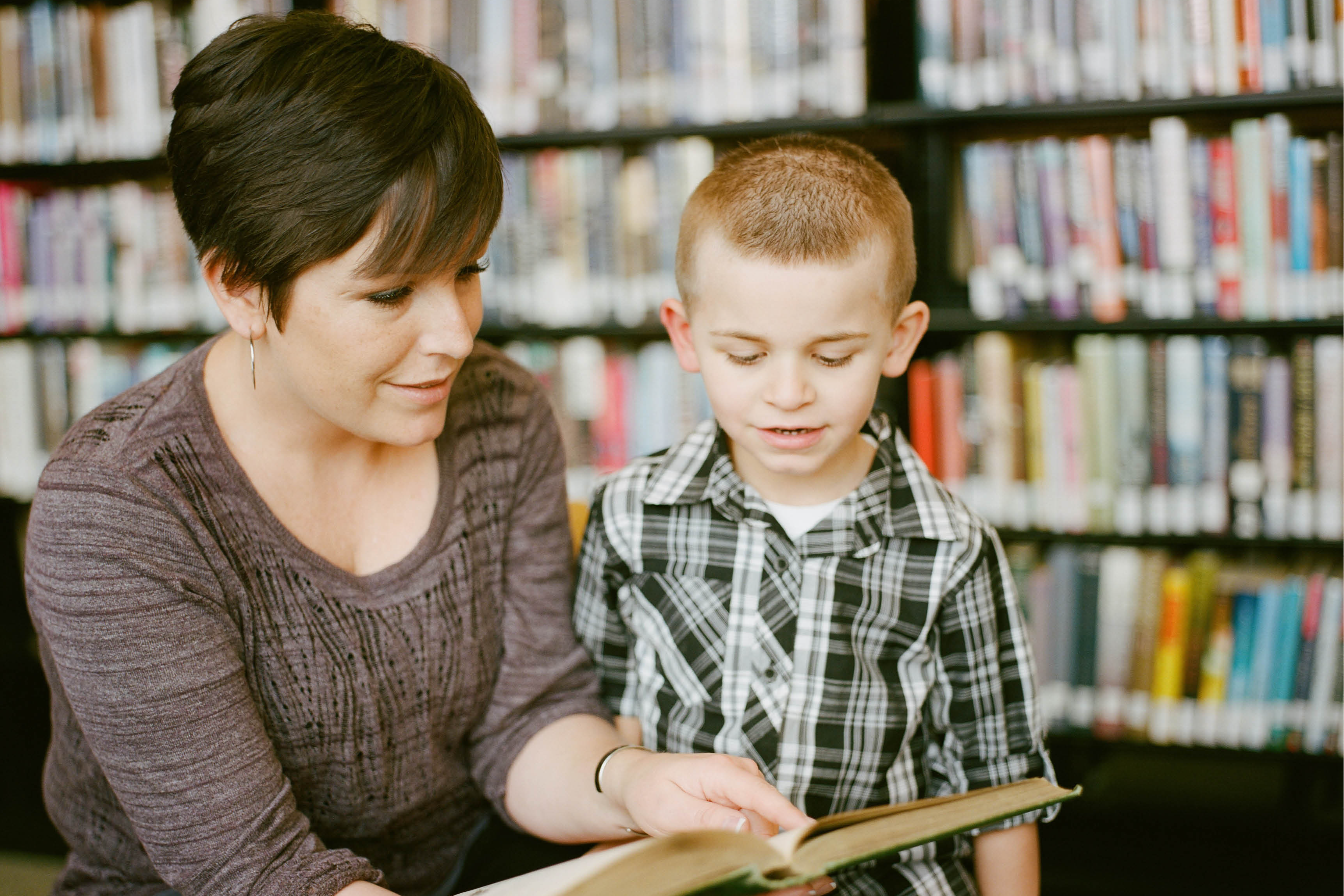 Woman and young boy reading a book in the library in front of a shelf of books blurry in the background. 