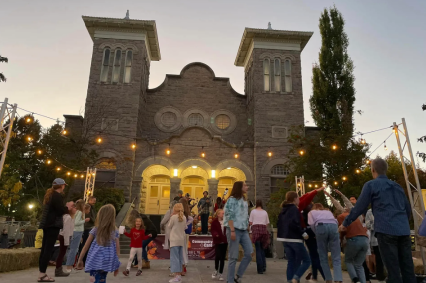 People dancing at dusk in front of the Tabernacle. a band plays on the steps and lights are strung above the crowd. 