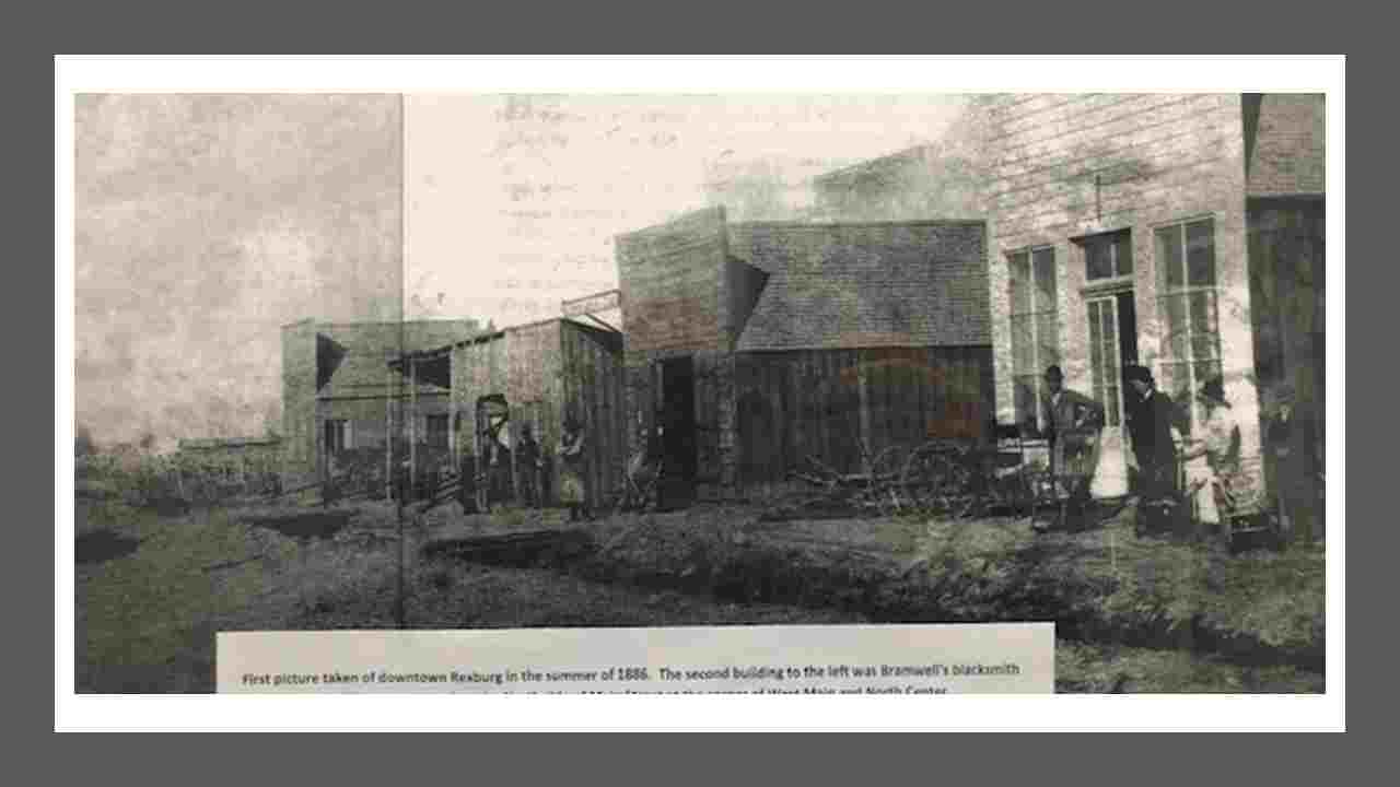 Historic photo of downtown Rexburg, noted as taken in 1886. 4 wood frame buildings on a dirt road with people in pioneer dress out front. 