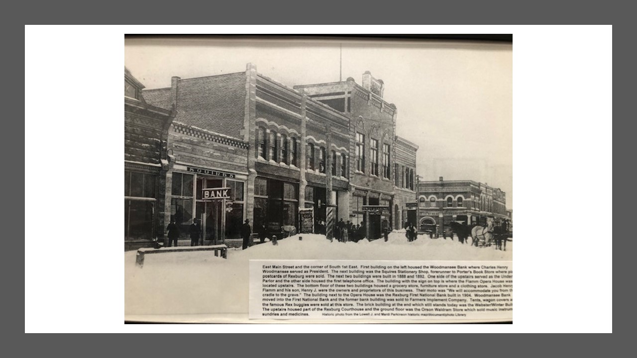 Historic photo of downtown Rexburg taken in the winter with snow covering the ground. A bank is visible  along with other connected buildings. Caption begins "East Main Street and the corner of S 1st E"