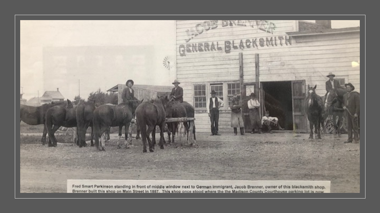 historic photo of a blacksmith shop and horses tethered to a post with men facing camera in late 19th c. period clothes.