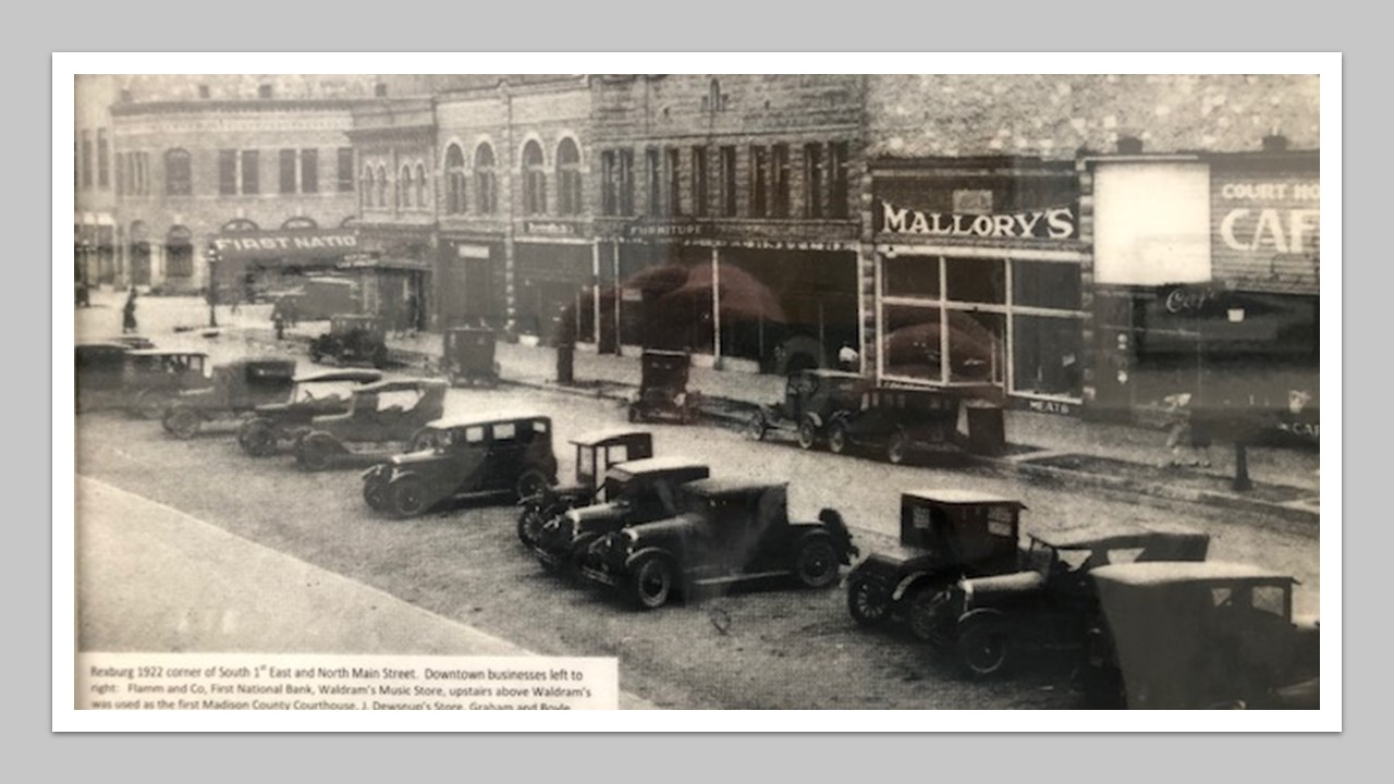 1920s cars, side-parked on Main street in front of the row of shops  and storefronts.