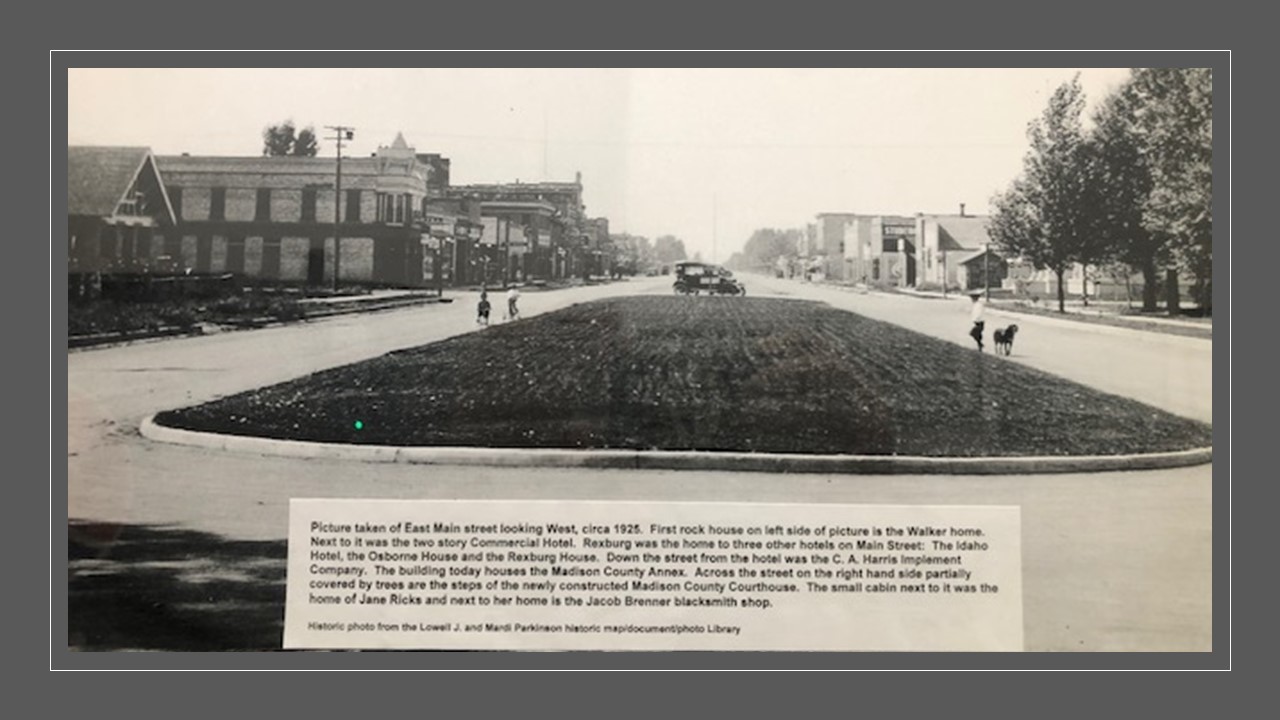 Historic photo of a wide green divider patch in the middle of Main street looking West with buildings and trees on the distant left and right and people walking on the street. A caption on the photo talks about buildings down the street including 3 hotels, a blacksmith,  the courthouse, and homes.