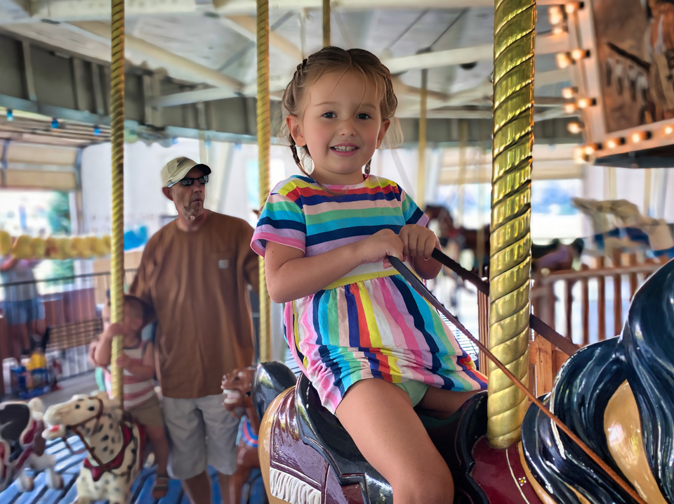 Girl on a carousel horse with older man visible standing in row behind looking away