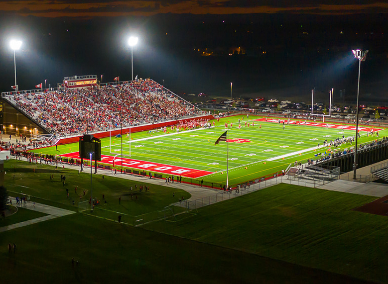 aerial photo of Bobcat Stadium at MHS at night, lit up by bright field lights, with fans packed into the stadium and players on the field. Photo by Cheney Photography