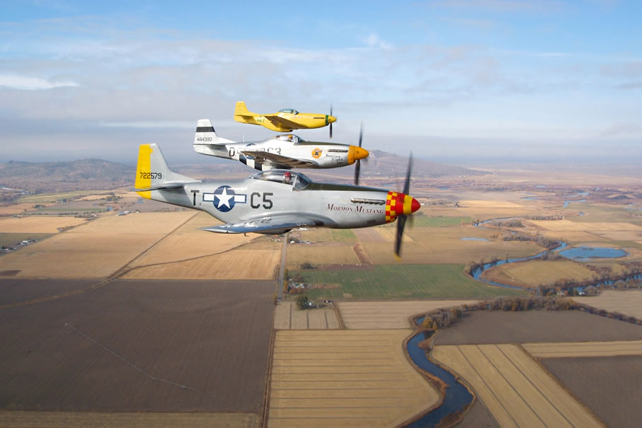 three small military planes in a row, flying in the air above the fields and river down below