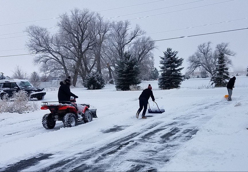 People plowing and shoveling snow in a long snow-covered driveway on a cloudy day