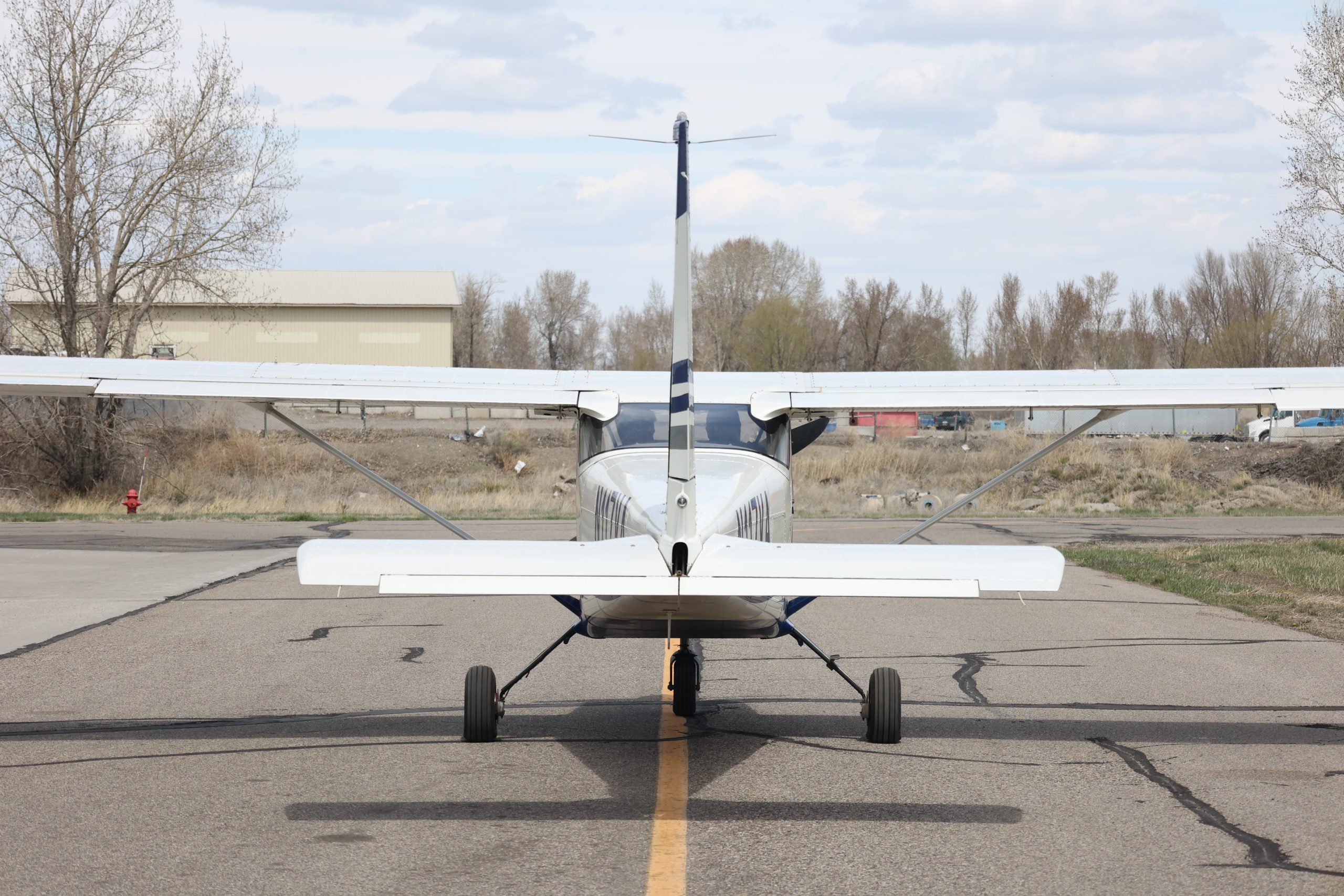 Small white airplane parked on a runway at an airport. Trees and buildings are visible in the background.