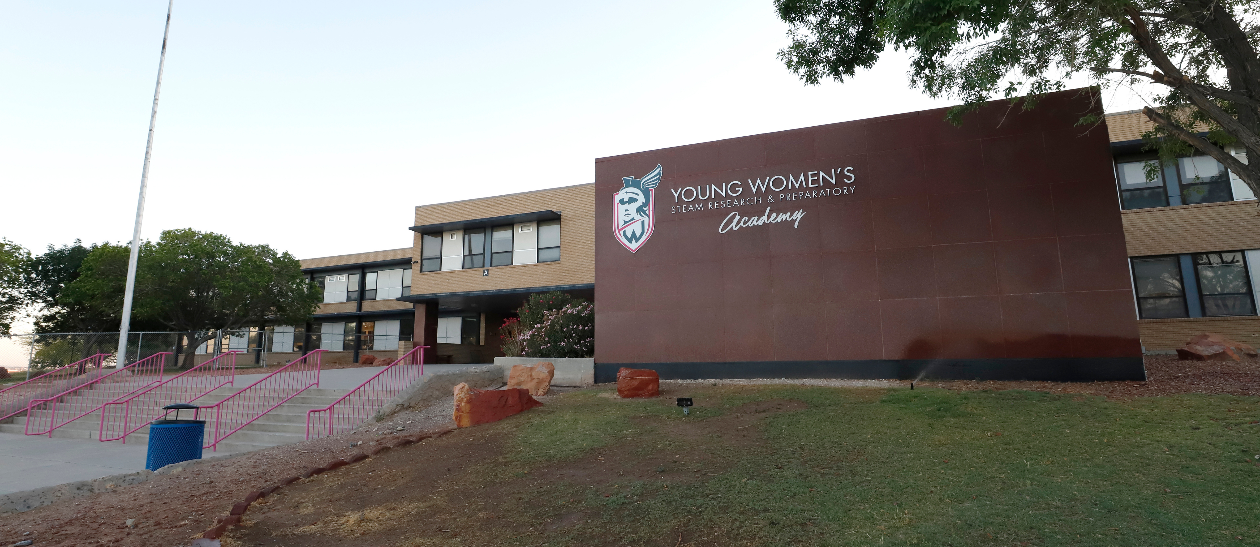 Building with brown sign, "Young Women's" and logo. Pink railings and grassy area in front.