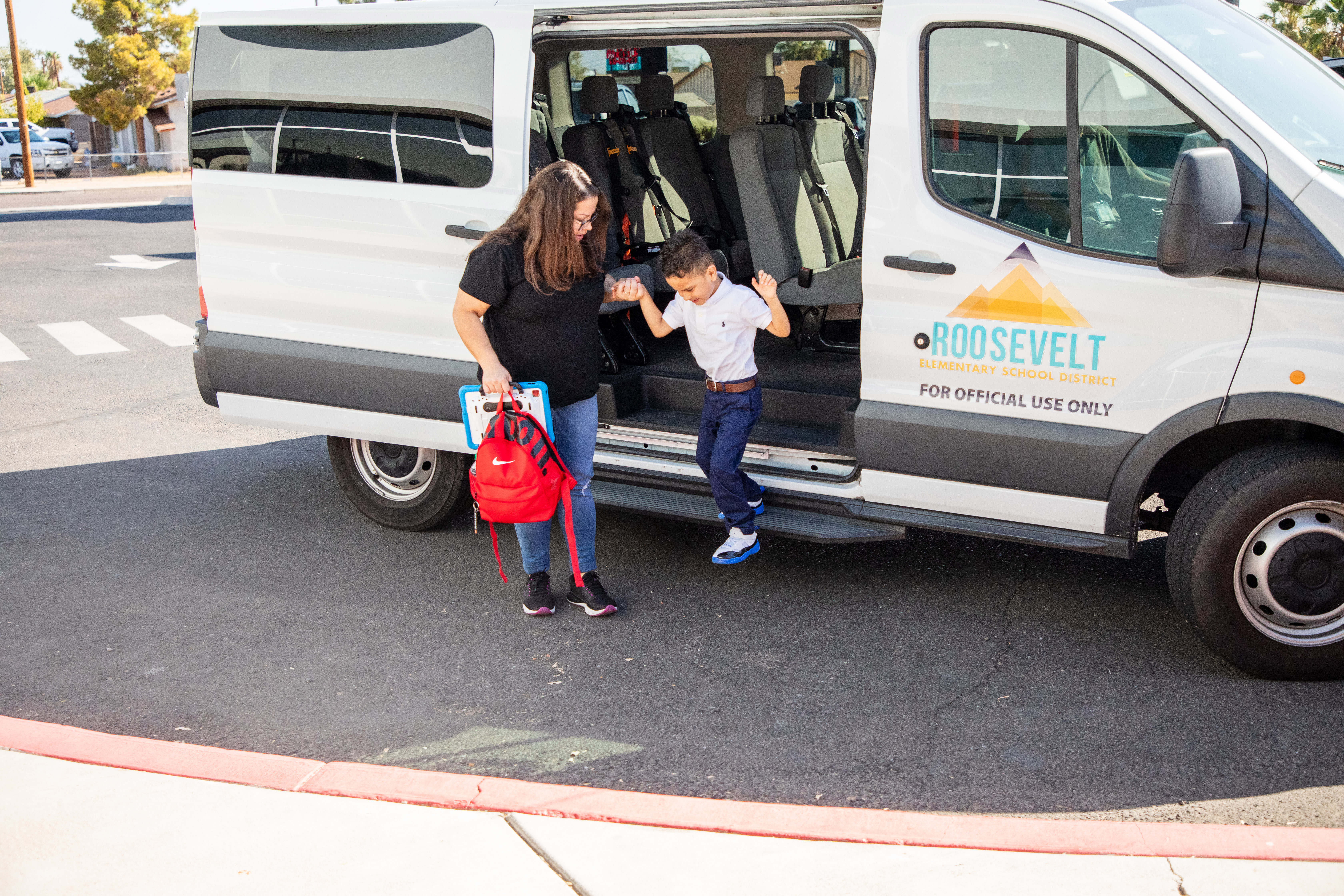 A young male student wearing a white shirt and navy pants being helped out of Roosevelt School District transportation by a staff member.