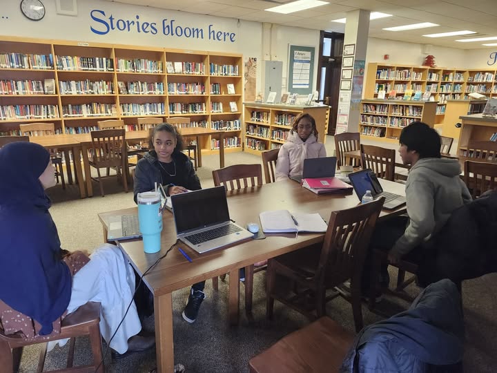 Students seated around a table working on laptops in a school library. Bookshelves line the room, and a wall sign reads “Stories bloom here