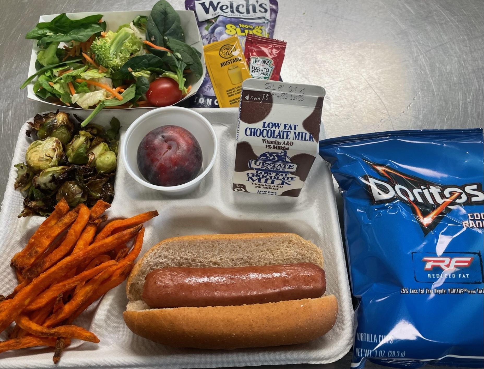 High School Lunch served 10.10.2024   Hot Dog, Sweet Potatoes Fries, Chili Roasted Brussels Sprouts, Side Salad, Chips, Apple, Chocolate Milk & Welch's Grape Slushy