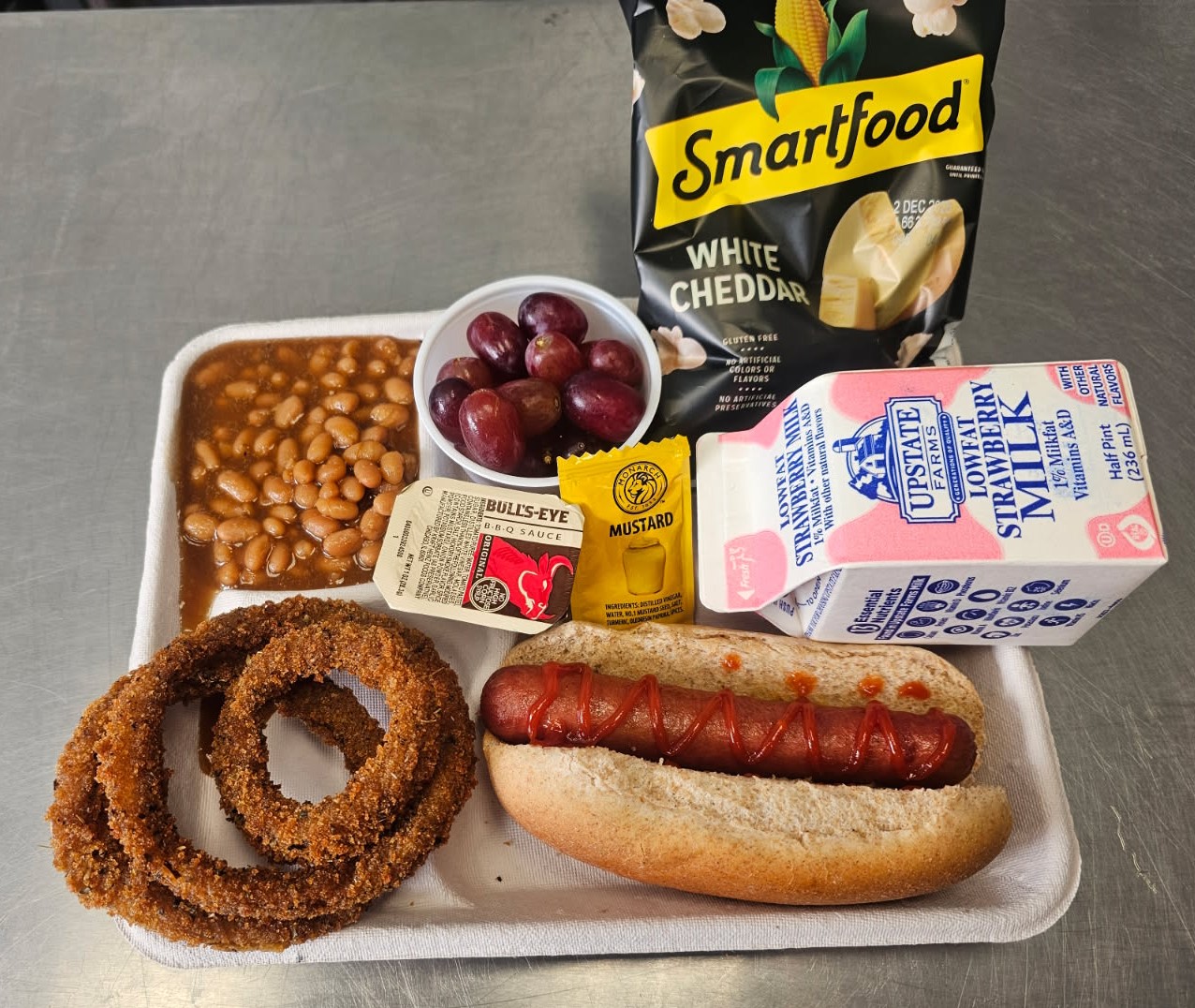 School #18 - Lunch served on 10.1.2025 Hot Dog, Onion Rings, Baked beans, Grapes, Cheddar popcorn & Strawberry milk