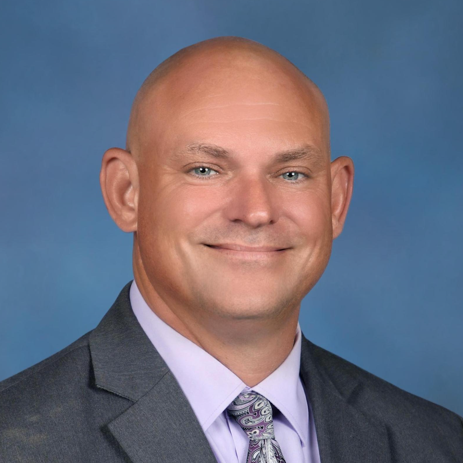 A man, bald, wearing a gray suit, purple shirt, and purple tie with white patterns. He smiles. Light blue background.