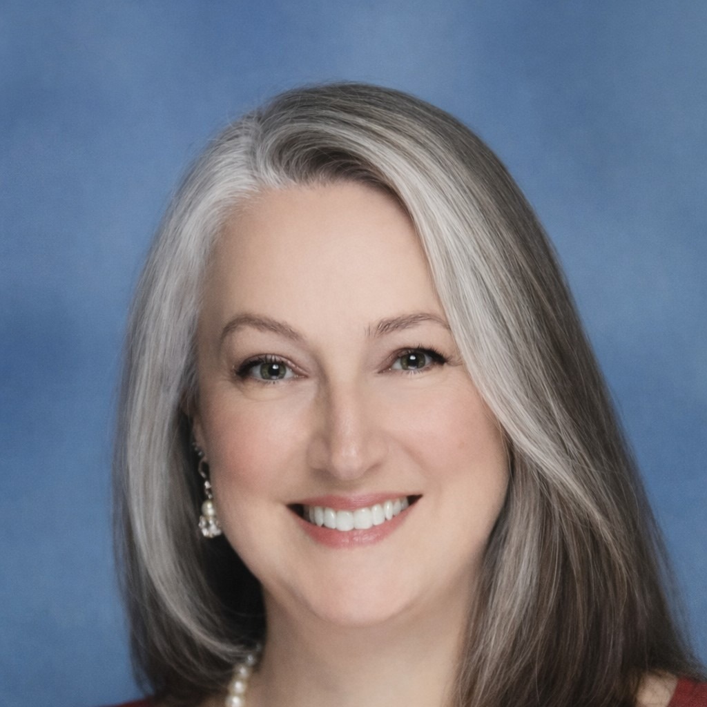 A woman with gray hair, wearing a red cardigan and a white pearl necklace, smiling for a portrait.