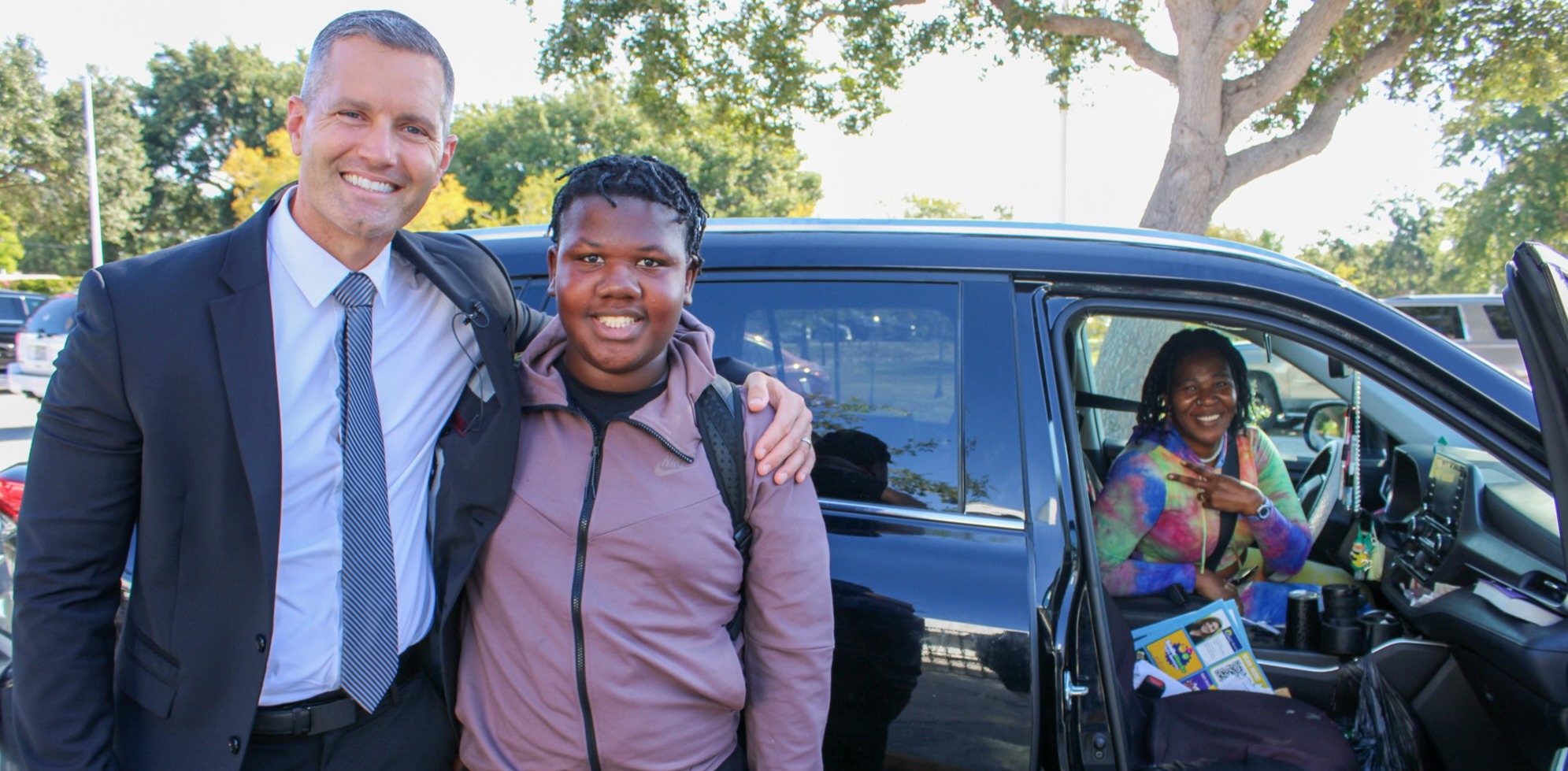 A man and a woman posing in front of a car, with the man wearing a suit and tie. The scene seems to be outside, under natural light.