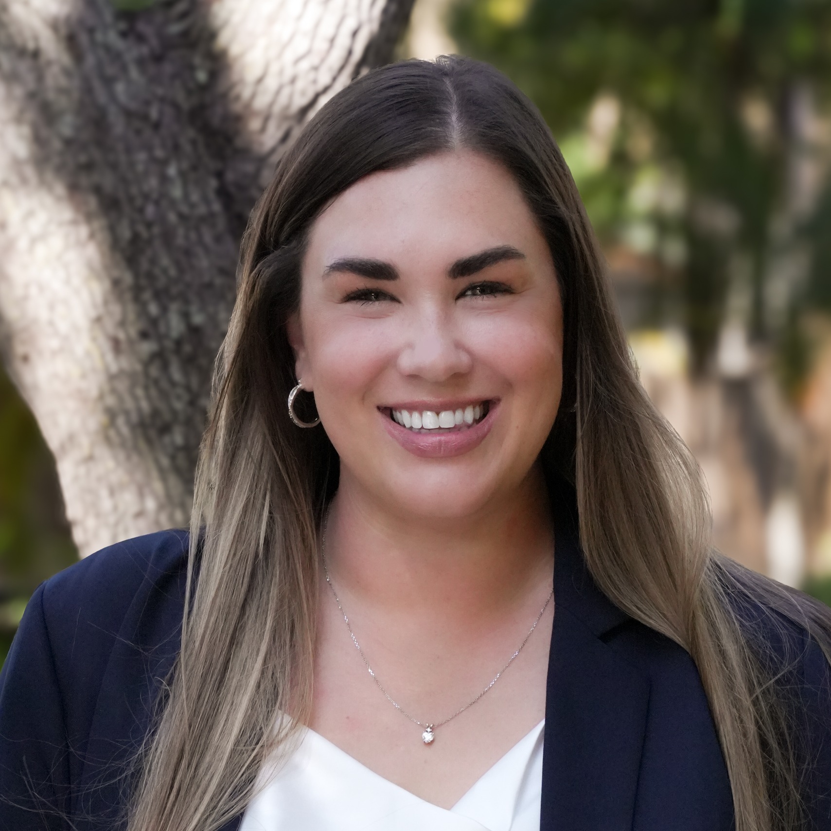 A smiling woman with long hair wearing a white blouse and blue blazer, standing outdoors with a tree in the background.
