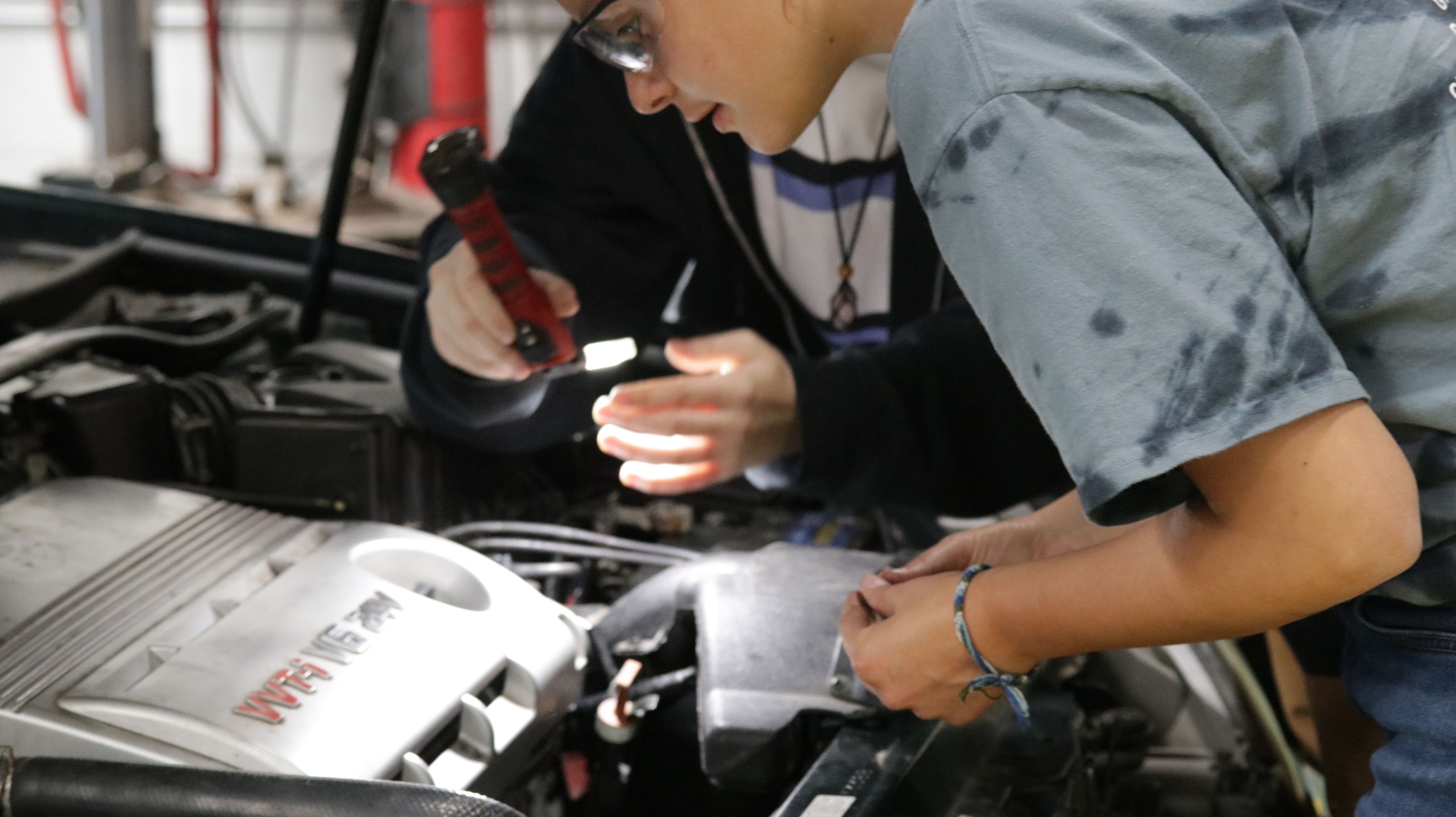 Two individuals working on a car engine, using tools and a flashlight