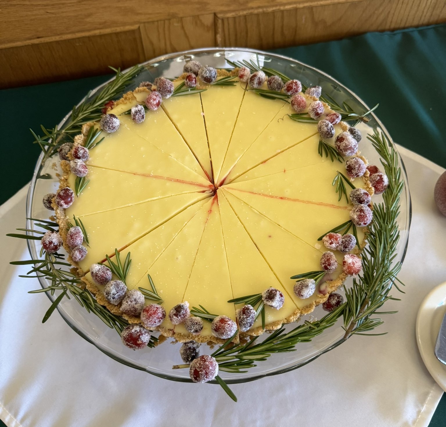 The image shows a cheesecake on a glass plate. The cheesecake is a light brown color and has a thick graham cracker crust. It is topped with whole sugared cranberries and a sprig of fresh rosemary.