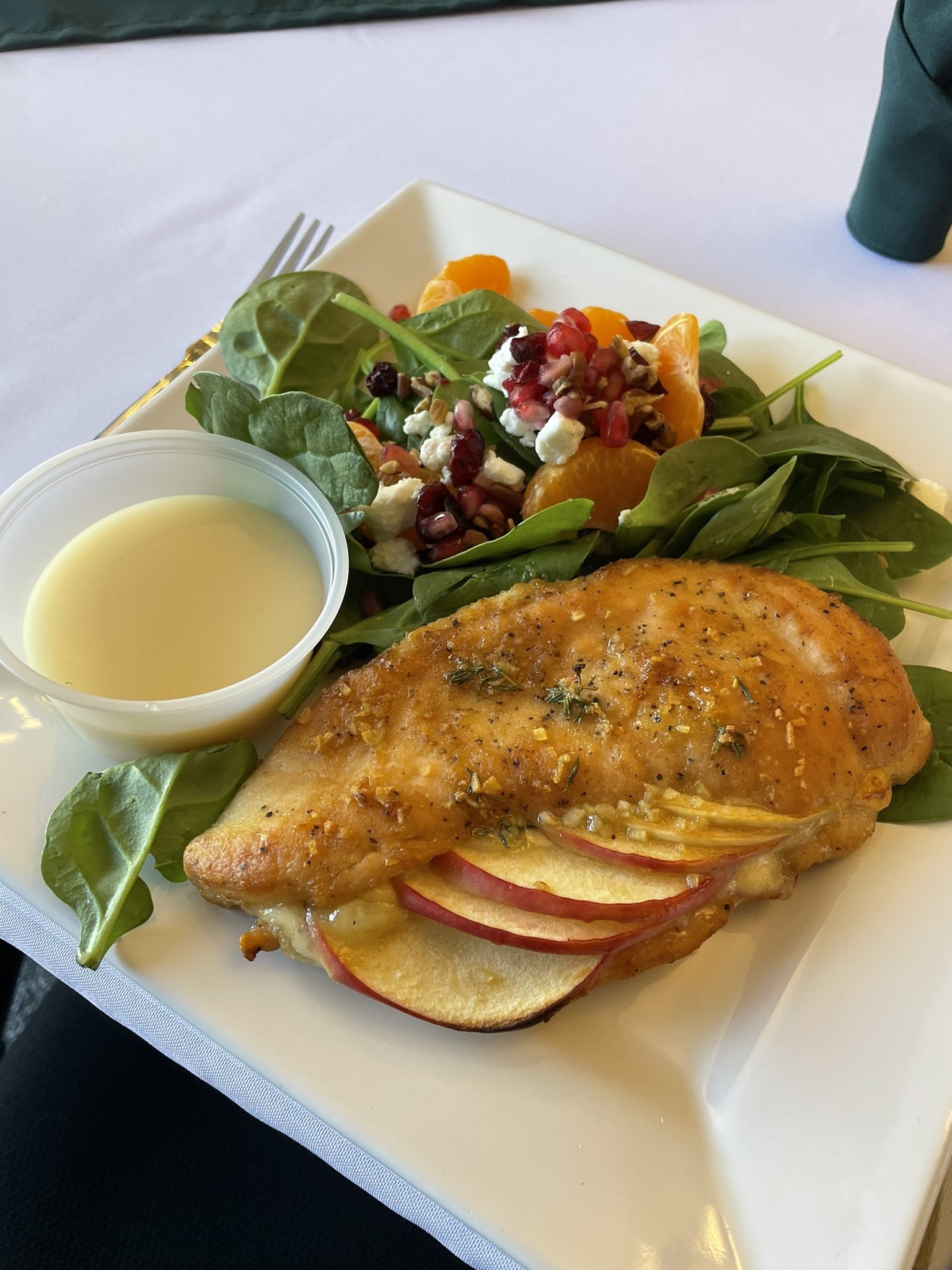 The image shows a white plate with a chicken breast and a salad. The chicken breast is cooked and sliced. The salad appears to have green leafy vegetables, possibly spinach, and sliced red apples. There is also a yellow lemon wedge on the plate.