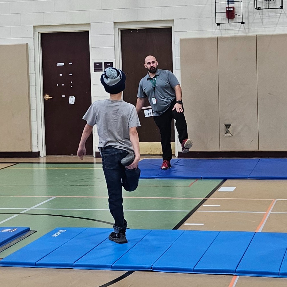 A man is observing another individual walking on blue mats laid out on a gymnasium floor.