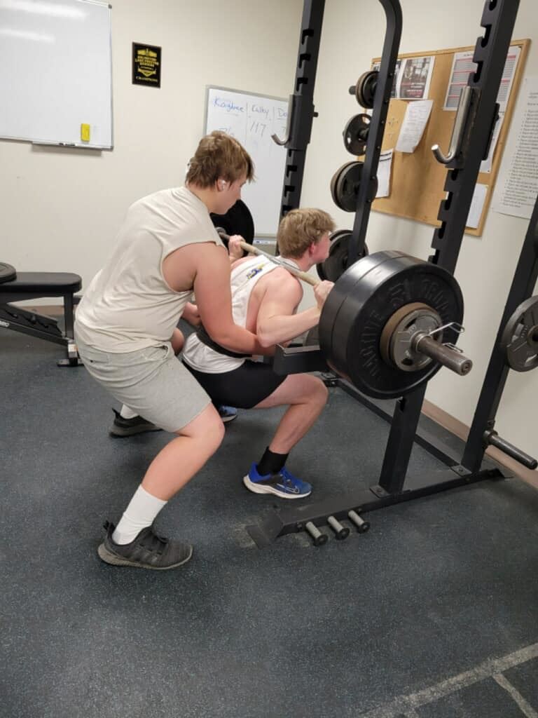 Two men performing squats in a gym, lifting a barbell.