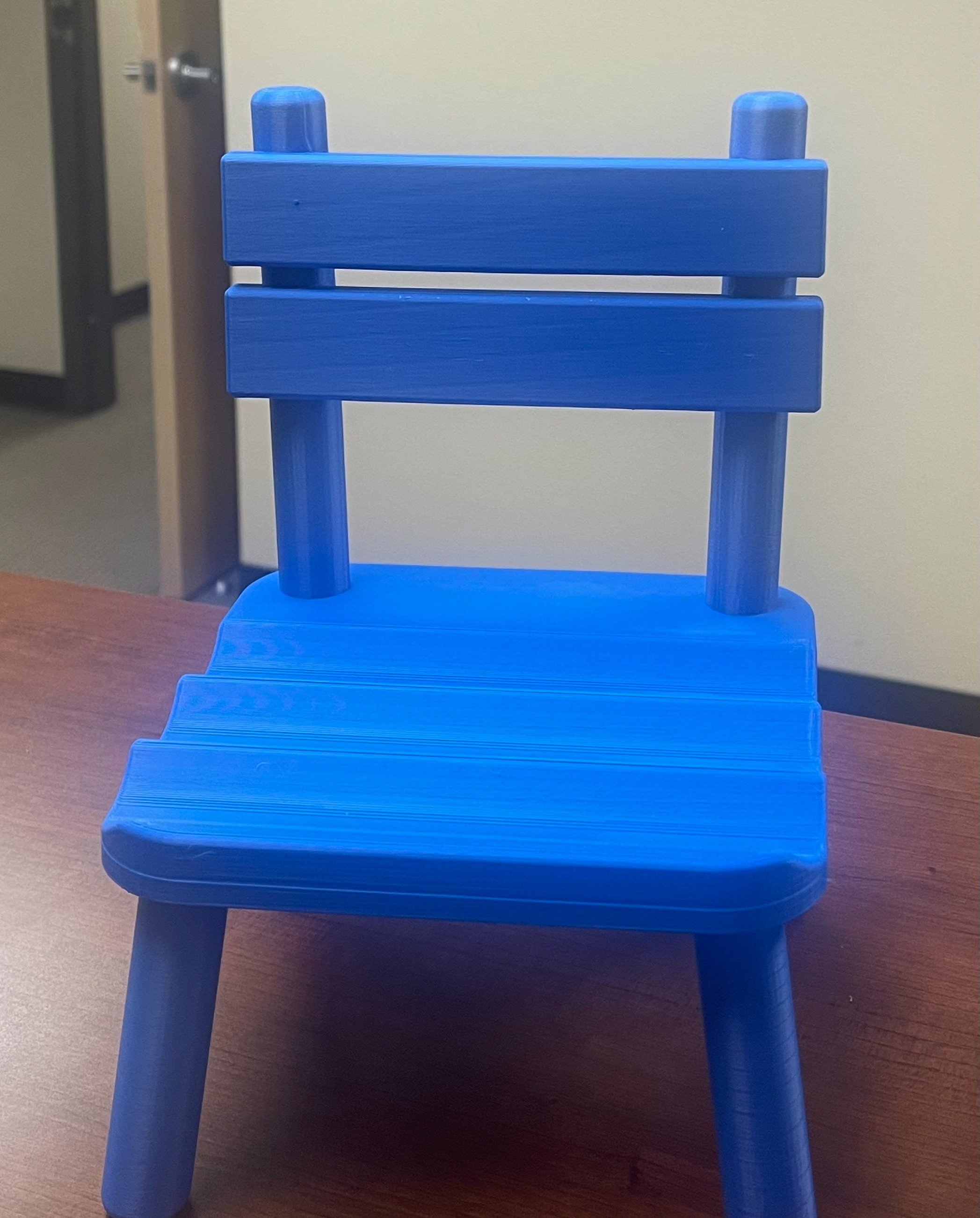 A blue plastic child's chair stands on a brown desk. Behind it is a door to a room.