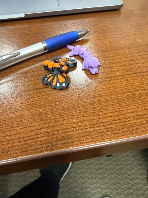 Wooden desk holds a blue pen, an orange butterfly charm, and a purple toy figure.
