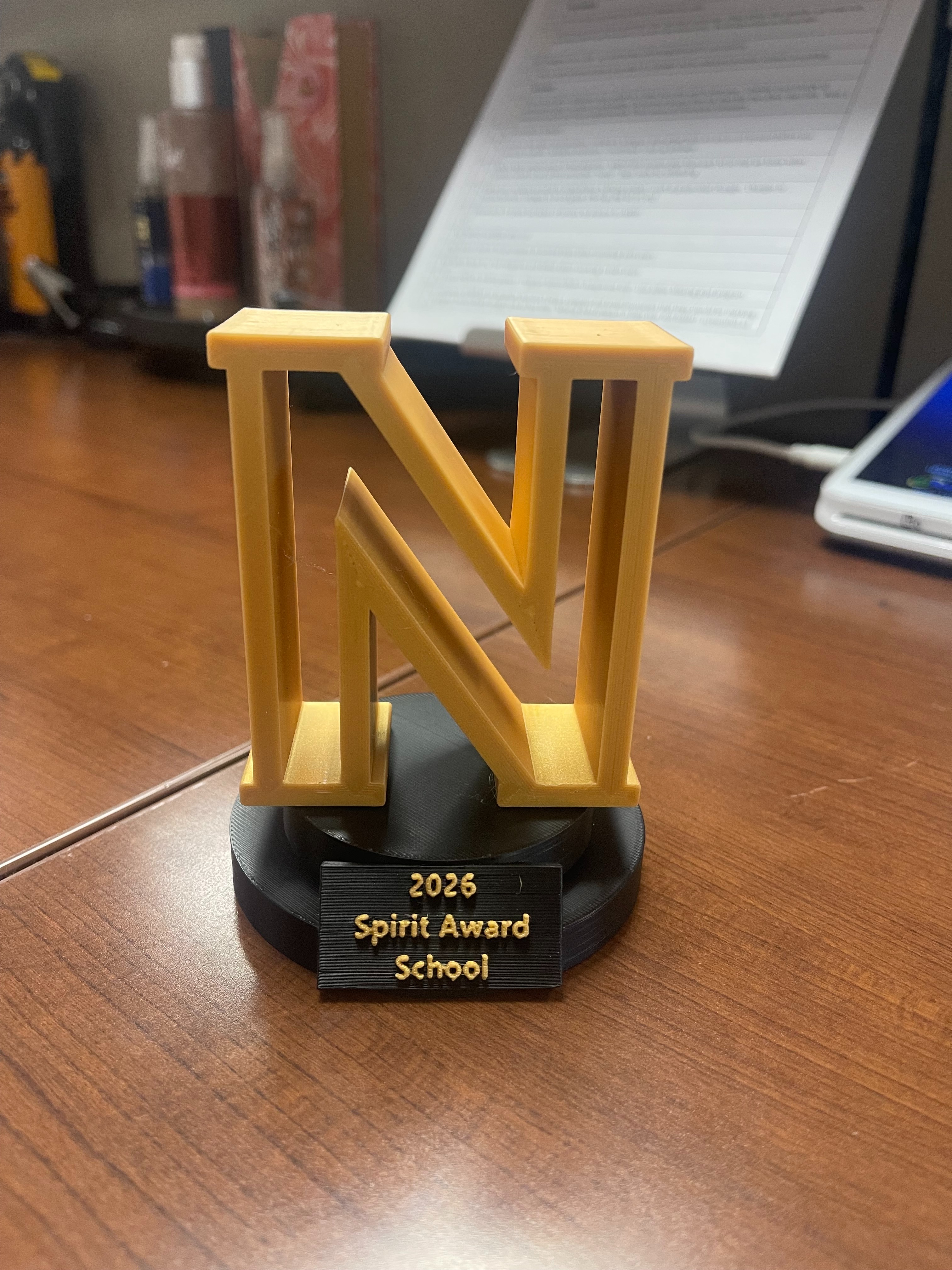 A wooden desk holds a golden trophy with the letters "N" and "V" standing vertically. Behind it, a music stand holds paper and a tablet.