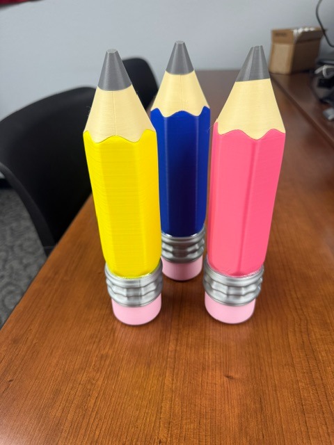 Three colored pencils, yellow, blue, and pink, stand upright on a wooden table. A black chair is behind the table.