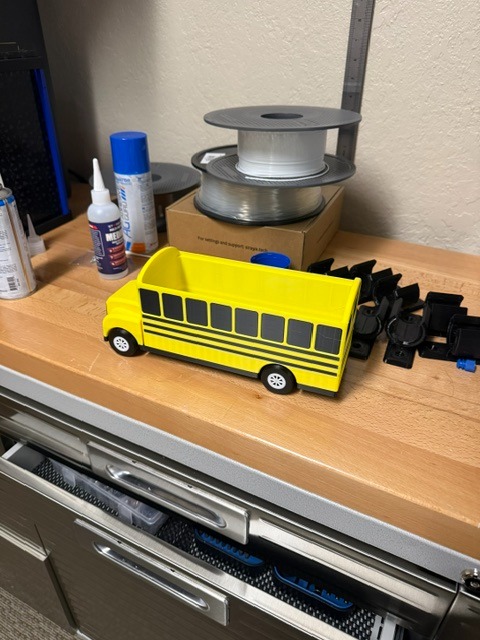 A yellow toy school bus on a wooden counter with various items, including a spool of wire, bottles, and a box.