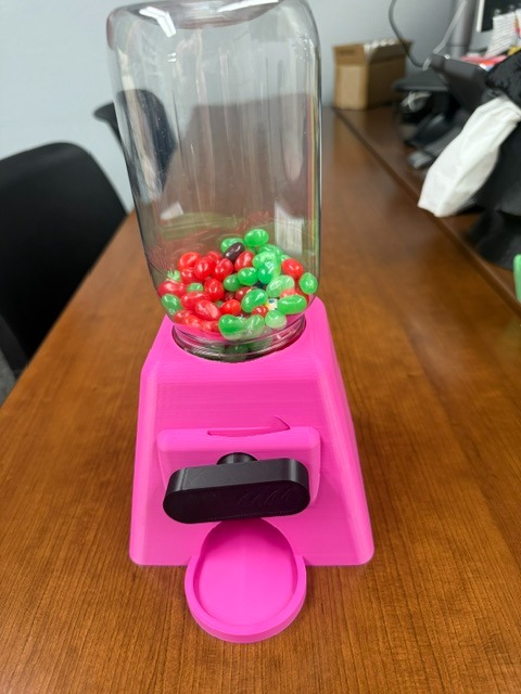 A pink candy dispenser with a transparent container full of multicolored candies sits on a wooden desk.