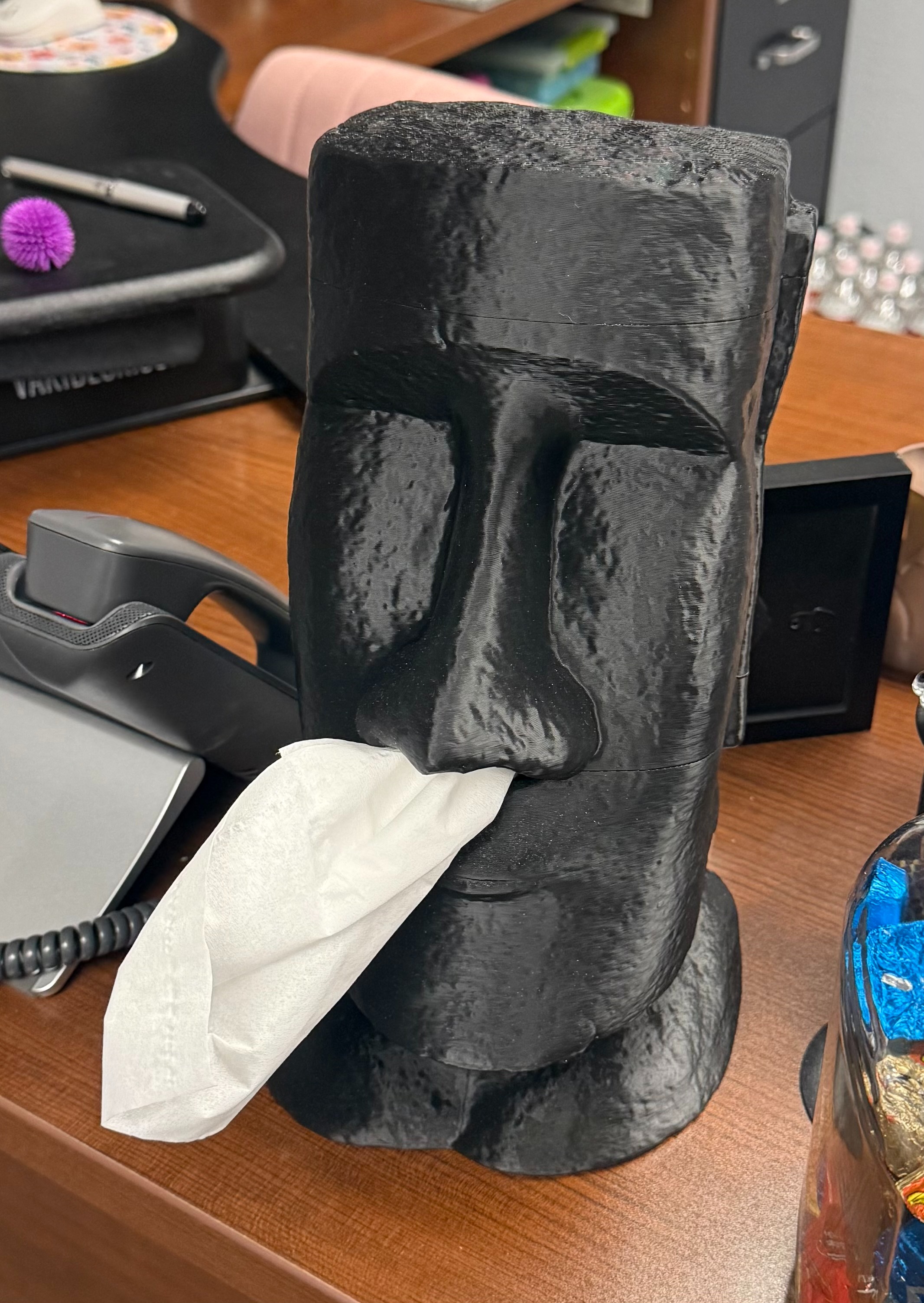 A black moai head sculpture sits on a desk with tissue in its mouth. Background includes a computer, pen, and office items.